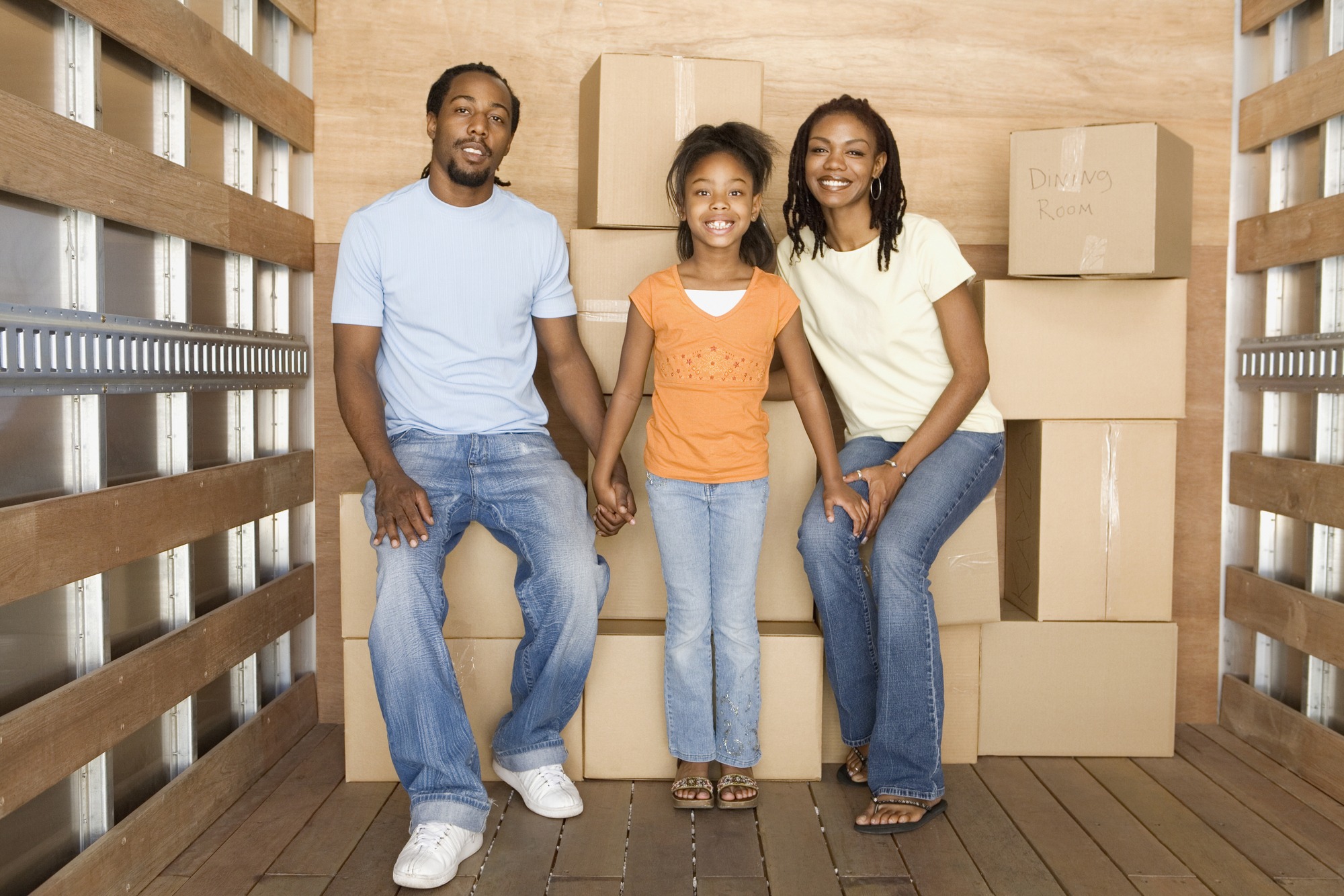 A happy family of three sits together on a box in a moving truck, surrounded by stacked cardboard boxes, symbolizing a new beginning.