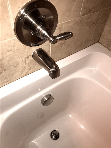 Modern bathroom faucet and tub controls against tiled wall, featuring a sleek design with a pull-up drain stopper and polished chrome finish.