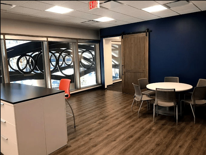 Modern office space featuring large windows, a round table with chairs, and a sliding door. Bright and functional, ideal for collaboration.