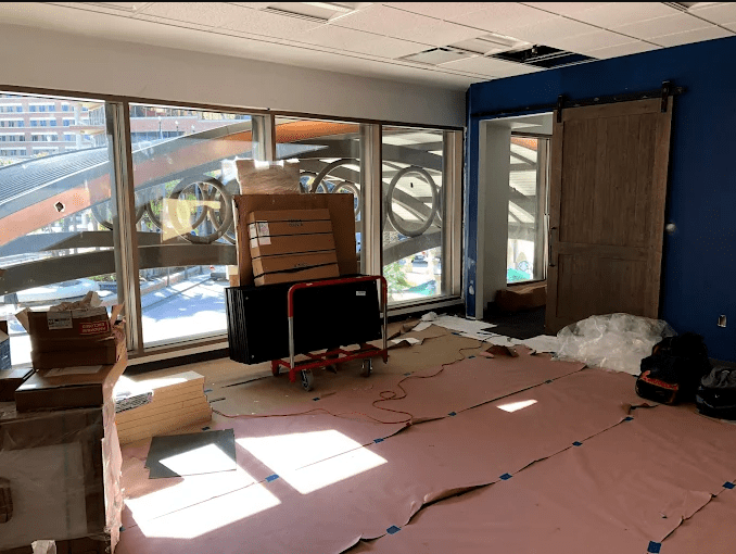 A partially furnished office space under renovation, featuring stacked boxes, a rolling cart, and exposed ceiling areas, with large windows allowing natural light.