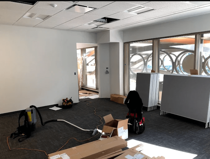 Renovation work in a bright, empty office space with cleaning equipment, boxes, and partitions arranged around large windows.