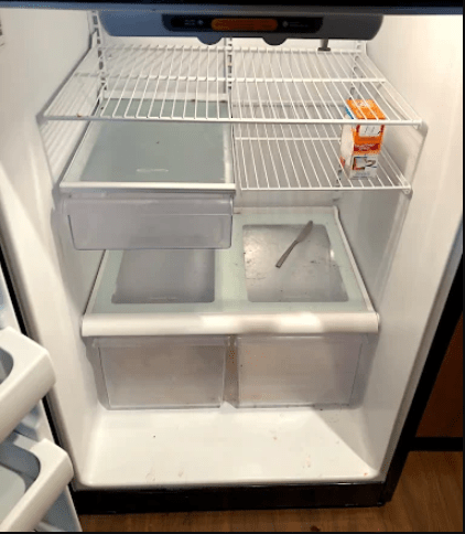 An empty refrigerator showing its interior shelves, a single juice box on the top shelf, and a spoon resting in the middle compartment.