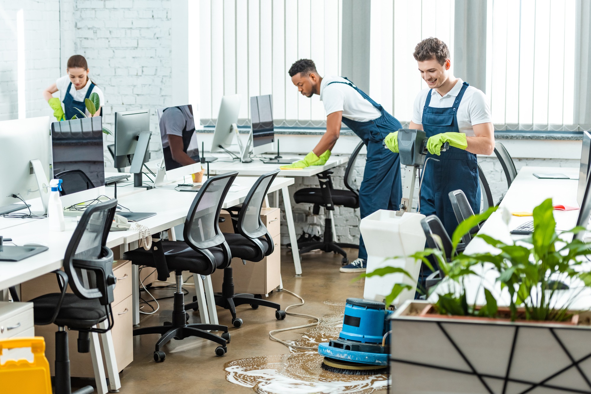 Cleaning crew in an office space, with two workers using cleaning equipment and another dusting surfaces, ensuring a tidy work environment.