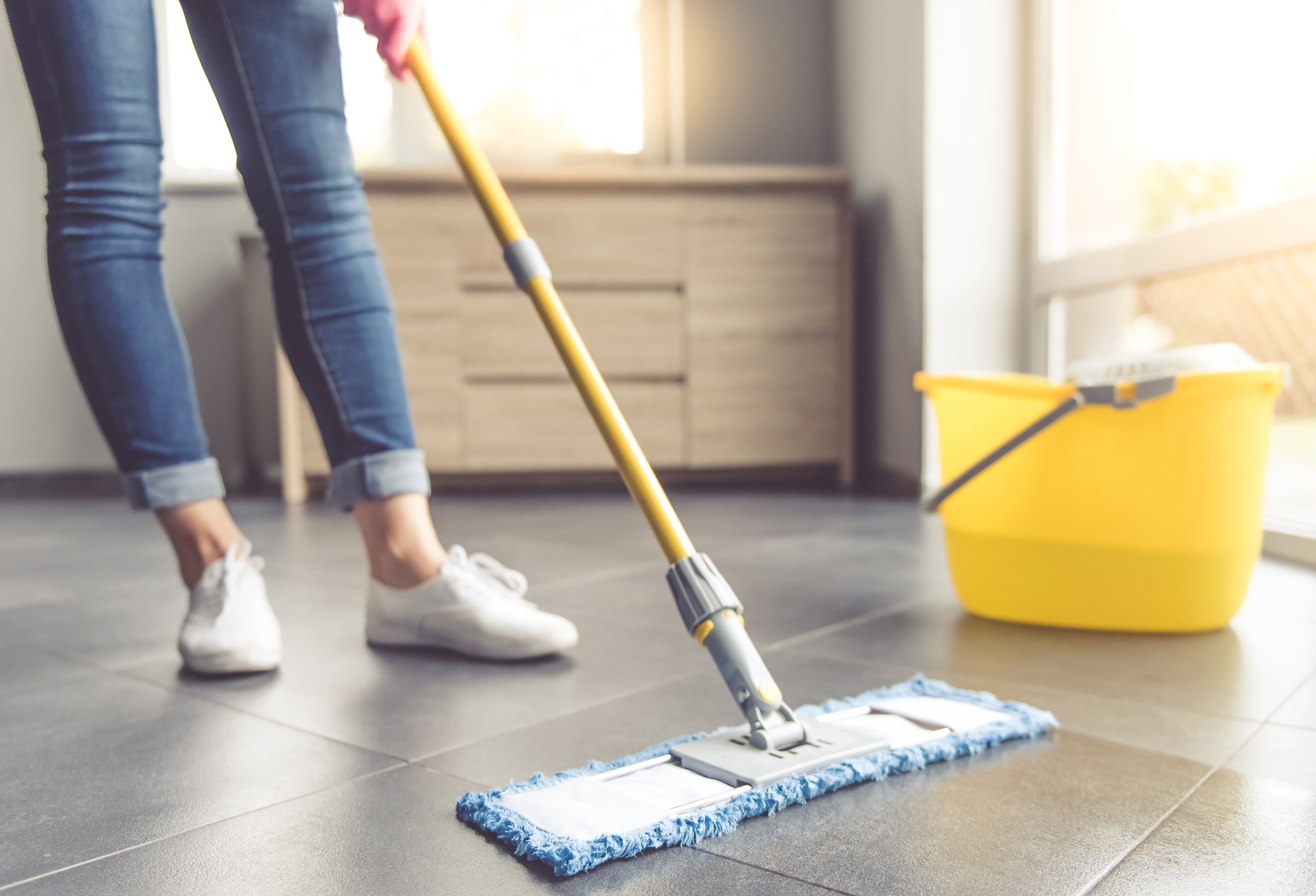 Person cleaning a tiled floor with a mop, wearing jeans and white shoes, with a yellow bucket nearby. Ideal for home cleaning tips.